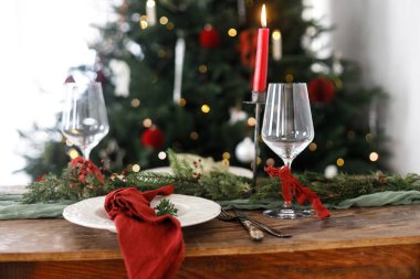 Modern tabaklar, kırmızı ve yeşil peçeteler, çatal bıçak, cam, köknar dalları, böğürtlen ve arkaplandaki Noel ağacındaki mumlarla süslü bir Noel sofrası. Mutlu Noeller ve mutlu tatiller!