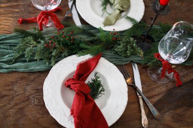 Aile yemeği için şık bir Noel sofrası. Peçete, çatal bıçak takımı, cam kaşık, kurdele, yeşil köknar dalları, ahşap masada duran kırmızı böğürtlenler. Noel Tatil Zamanı