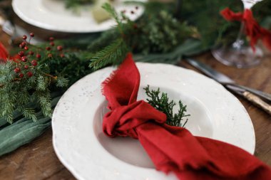 Mutlu Noeller! Noel aile yemeği için şık bir masa. Peçete, cam kaplama, yeşil ve kırmızı kurdeleler, köknar dalları ve kırsal masada mum bulunan modern tabak ve çatal bıçak takımı.