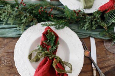 Aile yemeği için şık bir Noel sofrası. Peçete, çatal bıçak takımı, cam kaşık, kurdele, yeşil köknar dalları, ahşap masada duran kırmızı böğürtlenler. Noel Tatil Zamanı