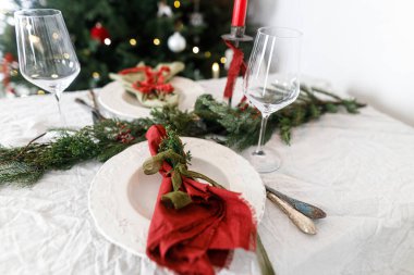 Aile yemeği için şık bir Noel sofrası. İçinde peçeteler, çatal bıçak takımı, cam kaşık, kurdele, yeşil köknar dalları, kırmızı böğürtlen ve keten masa örtüsü üzerine mum olan zarif bir tabak. Tatil zamanı.