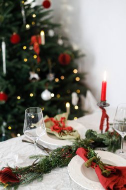 Modern tabaklar, kırmızı ve yeşil peçeteler, çatal bıçak, cam, köknar dalları, böğürtlen ve arkaplandaki Noel ağacındaki mumlarla süslü bir Noel sofrası. Mutlu Noeller ve mutlu tatiller!