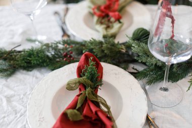 Aile yemeği için şık bir Noel sofrası. İçinde peçeteler, çatal bıçak takımı, cam kaşık, kurdele, yeşil köknar dalları, kırmızı böğürtlen ve keten masa örtüsü üzerine mum olan zarif bir tabak. Tatil zamanı.
