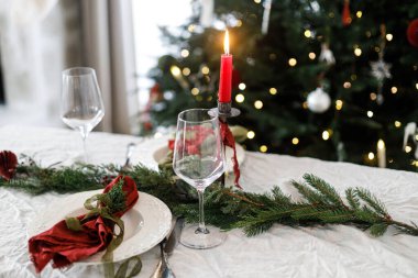 Aile yemeği için şık bir Noel sofrası. İçinde peçeteler, çatal bıçak takımı, cam kaşık, kurdele, yeşil köknar dalları, kırmızı böğürtlen ve keten masa örtüsü üzerine mum olan zarif bir tabak. Tatil zamanı.
