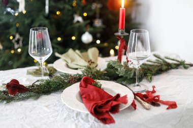 Aile yemeği için şık bir Noel sofrası. İçinde peçeteler, çatal bıçak takımı, cam kaşık, kurdele, yeşil köknar dalları, kırmızı böğürtlen ve keten masa örtüsü üzerine mum olan zarif bir tabak. Tatil zamanı.