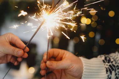 Noel ağacı ve şömine karşısında havai fişek tutan eller süslü, rahat bir oturma odasında. Mutlu yıllar! Atmosferik aile kutlaması, ellerinde yanan maytaplarla.