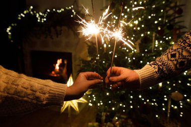 Akşam sıcak oturma odasında Noel ağacı ışıklarına karşı havai fişek tutan eller. Yeni yılınız kutlu olsun! Atmosferik aile kutlaması, ellerinde yanan maytaplarla.