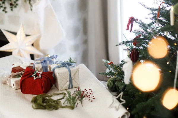 Mutlu Noeller ve mutlu tatiller! Paketlenmiş şık Noel hediyeleri ipek yaylar, yeşil dal ve böğürtlenler, kırsal masadaki süslere karşı dekore edilmiş yılbaşı ağaçları..