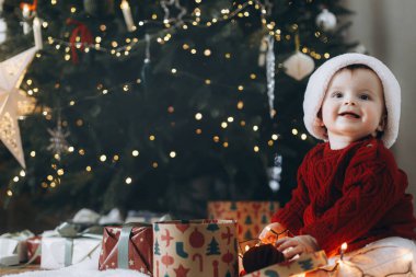 Mutlu Noeller ve mutlu tatiller! Noel Baba şapkalı şirin bebek şık hediye kutusuyla oynuyor ve arka planda süslü Noel ağacı süsleri var. Tatlı atmosferik an