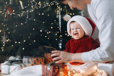 Noel Baba şapkalı şirin bebek modern Noel ağacının altında elinde çelenk ışıkları olan hediye kutularıyla annesine ağlıyor. Üzgün bir bebek. Tatil zamanı.