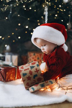 Mutlu Noeller ve mutlu tatiller! Noel Baba şapkalı şirin bebek şık hediye kutusuyla oynuyor ve arka planda süslü Noel ağacı süsleri var. Tatlı atmosferik an