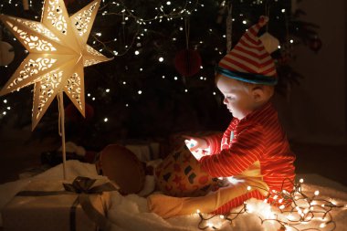 Mutlu Noeller ve mutlu tatiller! Noel kostümü giymiş şirin bir bebek hediye kutusunda oturmuş süslenmiş Noel ağacının arka planında sihirli bir ışık var. Atmosfer arifesi, sihirli an