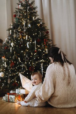 Mutlu Noeller ve mutlu tatiller! Anne sevimli bebeğe sarılıyor ve dekore edilmiş ışıklandırmalı ve hediyeli bir Noel ağacının arka planında oturuyor. Aile tatili zamanı. Atmosferik güzel bir an