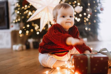Süslü süveter giymiş sevimli mutlu bir bebek şık hediyelerle oturuyor ve yıldızlı bir Noel ağacının arka planında çelenk ışıkları var. Atmosferik tatil zamanı, Mutlu Noeller!
