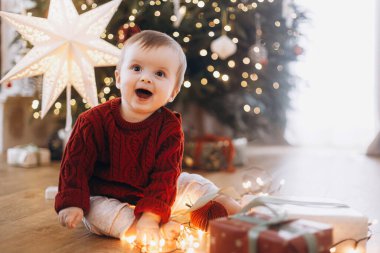 Süslü süveter giymiş sevimli mutlu bir bebek şık hediyelerle oturuyor ve yıldızlı bir Noel ağacının arka planında çelenk ışıkları var. Atmosferik tatil zamanı, Mutlu Noeller!
