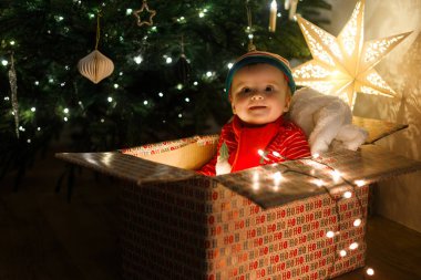 Noel kostümlü tatlı bir bebek. Hediye kutusunda oturuyor. Işıkları aydınlatılmış Noel ağacına ve akşamları yıldızlara karşı. Atmosfer arifesi, sihirli an. Mutlu Noeller ve mutlu tatiller!