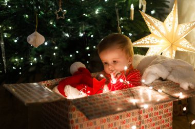 Mutlu Noeller ve mutlu tatiller! Noel Baba şapkalı şirin bebek büyük bir hediye kutusunda oturuyor. Arka planda çelenk ışıkları var. Noel ağacı ve ışıldayan yıldız. Atmosferik xmas arifesi
