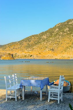 Beyaz boyalı ahşap sandalyeler ve çiçek desenli masa örtüsü. Gün batımında sahilin kenarında.