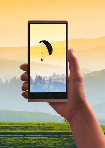 Paraglide silhouette in a mountains on a screen of smartphone taking by girl hand. Travel and sport concept.