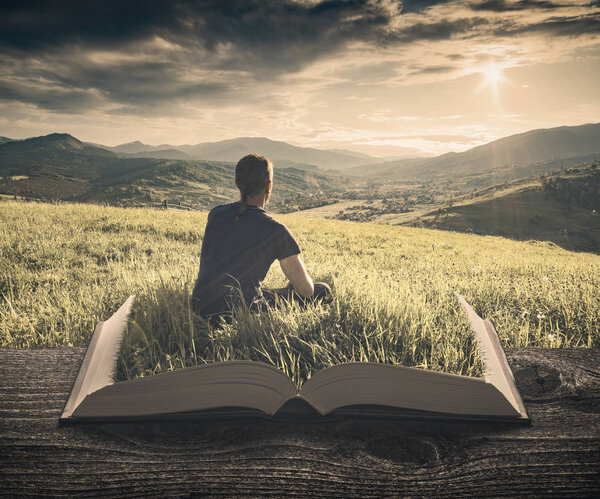 Man in a grass enjoy the valley on the pages of an open magical book. Majestic landscape. Travel and education concept. Vintage filter stylization.