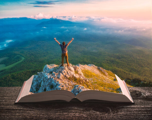 Young hiker man standing on a cliff above the mountain valley on the pages of an open magical book. Majestic landscape. Travel and education concept.