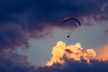 Gün batımında bulutlu bir gökyüzünde uçan paraglider silueti.