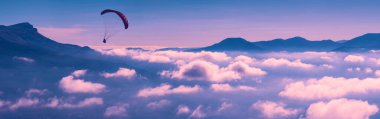 Bir dağ vadisinde bulutların üzerinde uçan paraglider silueti.