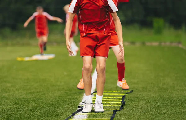 Young Athletes Training with Football Equipment. Football Speed Training with Ladder. Young Footballer Jumping in Red Sportswear at Training Session on Grass Soccer Field