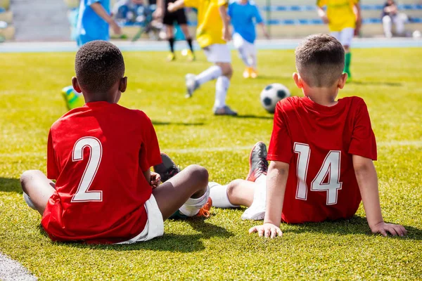 Futbol oyunu oynayan çocuklar çok ırklı grup. Futbol sahasında çim üzerinde oturan ve destek takım arkadaşları kırmızı futbol formaları giyen iki Genç Irklararası erkek — Stok fotoğraf