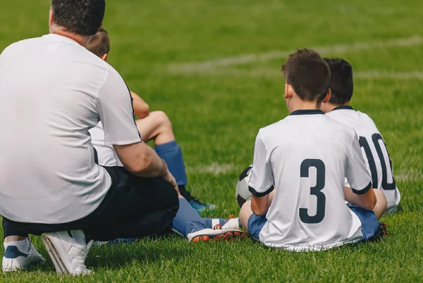 Fußballjungs sitzen mit Trainer auf dem Schulrasenrasenplatz. Trainer der Kinderfußballmannschaft im Gespräch mit jungen Spielern in der Halbzeitpause — Stockfoto