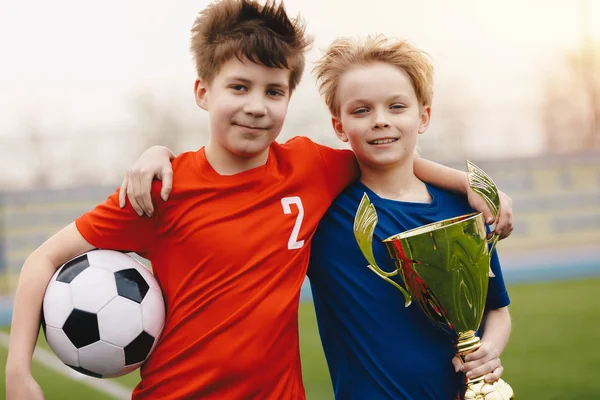 サッカーボールと黄金のトロフィーを持つ2人の幸せな少年サッカー選手。赤と青のジャージスポーツウェアでフィールド上の子供のサッカー選手 — ストック写真