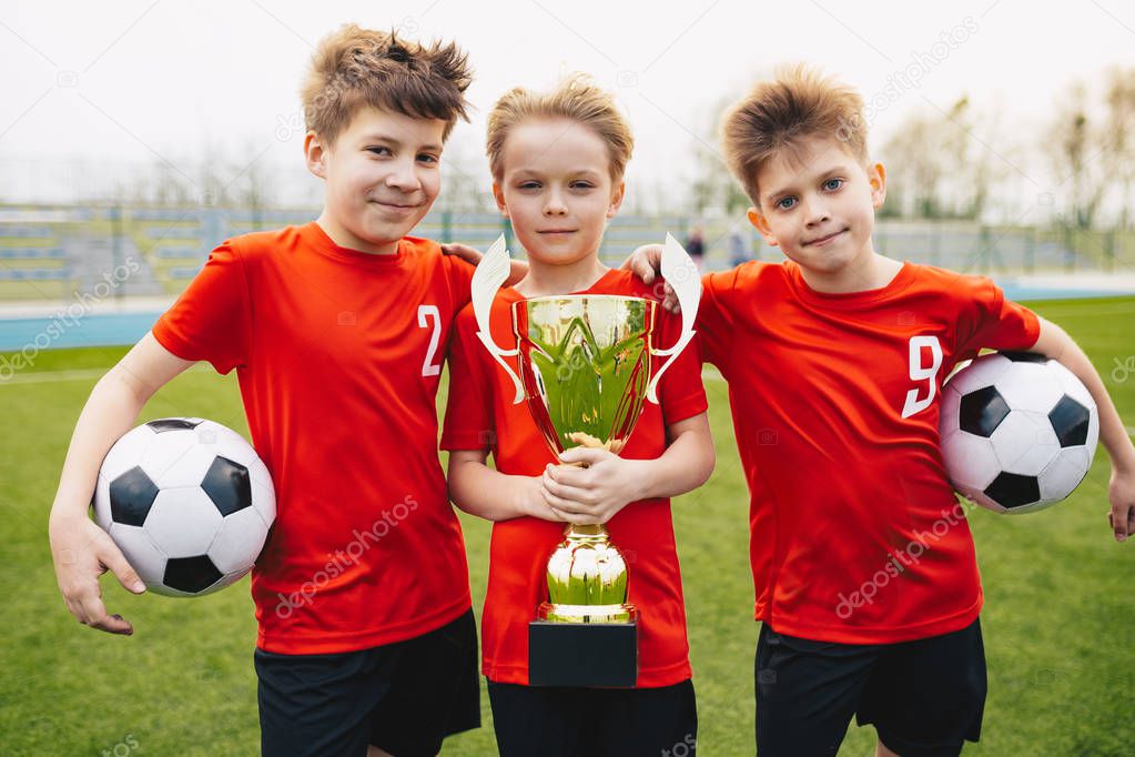 Preteen happy soccer players after final game. Boys holding golden cup and soccer balls. Sports portrait of three happy football players. Football youth junior team posing outdoor and facing camera