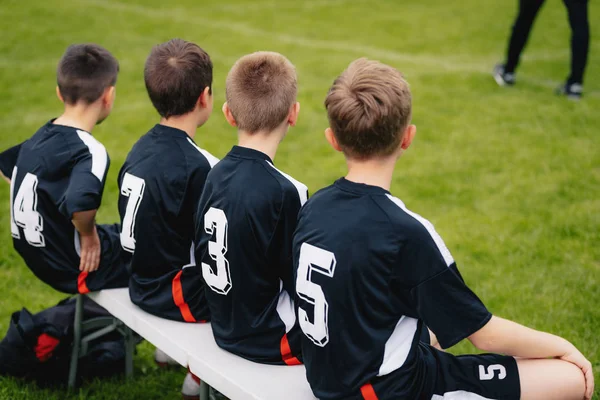 Meninos no banco de futebol durante o jogo de futebol. Treinador da equipe de esportes da juventude no fundo — Fotografia de Stock