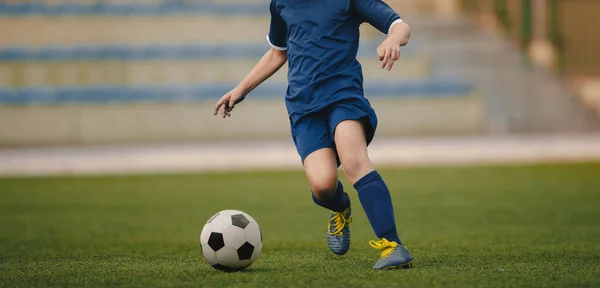 ジュニアレベルのサッカー選手がボールで走っています サッカークリートのサッカー選手と草のピッチ上の古典的なサッカーボールを蹴るジャージーブルーのユニフォーム 背景にあるスポーツアリーナ — ストック写真