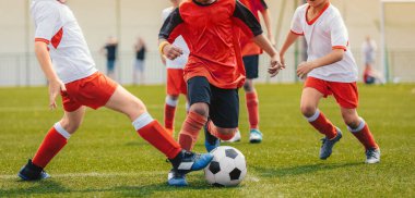 Futbol oynayan çok ırklı çocuklar. Genç çocuklar çim futbol sahasında futbol topunun peşinden koşuyorlar. Kırmızı ve beyaz formalı çocuklar.