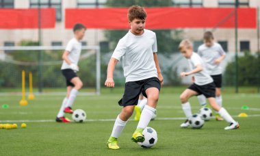 Çocuklar yapay çim antrenman sahasında futbol toplarını tekmeliyor. Genç çocuk top sürüyor ve top sürme becerilerini geliştiriyor. Beden eğitimi dersi, okul eğitimi sunumu.
