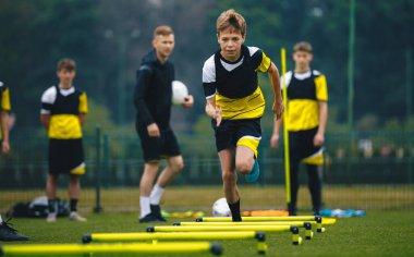 Çocuklar iki genç koçla futbol antrenmanında. Ortaokul futbolcusu antrenman sırasında engelli koşuyor ve atlama egzersizi yapıyor. Futbol antrenmanları çevikliği engelliyor