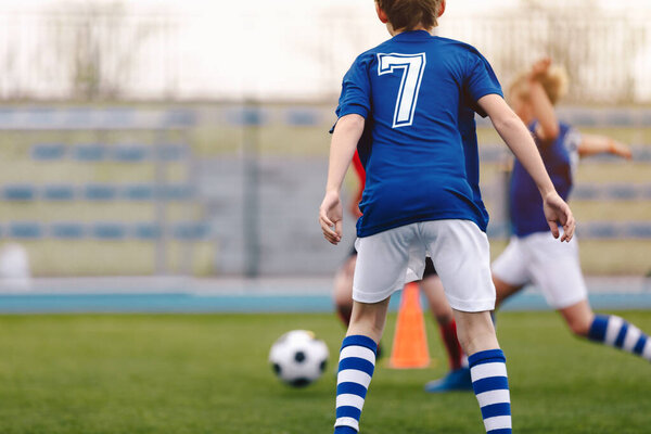 Back of young soccer boy playing game on grass school field. Young football player in blue jersey sports shirt with number seven on his back