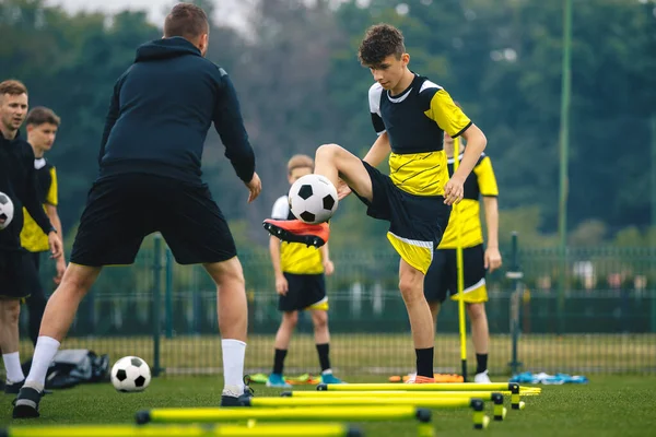 Teenagers on soccer training camp. Boys practice football witch young coaches. Junior level athletes improving soccer skills on outdoor training. Player kick soccer ball to coach and ladder skipping