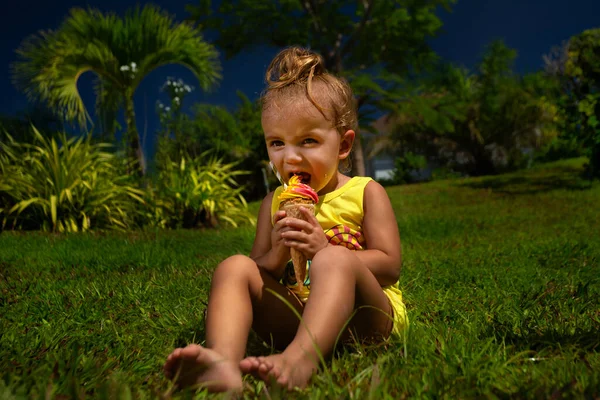 Comic portrait of Pretty baby hungry girl kid eating big ice cream in waffles cone, sittinng on grass in park in the summer tim