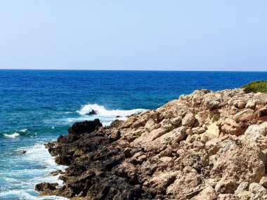 Resimli plaj, kumlu kıyıya vuran dalgalar. Deniz dalgaları kayalara çarpıyor. Kıbrıs adası, şemsiyeler, turist botları, Akdeniz.