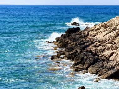 Resimli plaj, kumlu kıyıya vuran dalgalar. Deniz dalgaları kayalara çarpıyor. Kıbrıs adası, şemsiyeler, turist botları, Akdeniz.