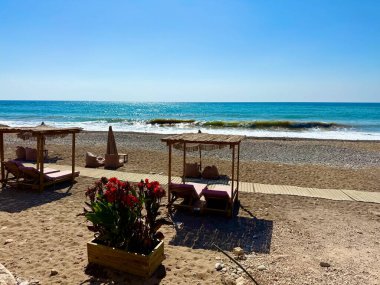 Halk plajı, yaz mevsimi, güzel güneşli hava. Resimli plaj, kumlu kıyıya vuran dalgalar. Deniz dalgaları kayalara çarpıyor. Kıbrıs adası, şemsiyeler, turist botları, Akdeniz.