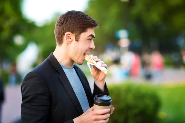 Closeup portrait of hipster man eating pastry and holding coffee at green park background.
