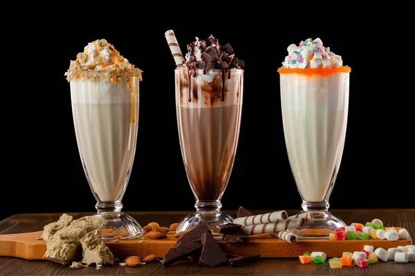 Three sweet milkshakes with chocolate, marshmallow, halva and whipped cream at a wooden board on a table isolated at black background.