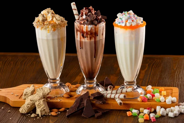 Three sweet milkshakes with chocolate, marshmallow, halva and whipped cream at a wooden board on a table isolated at black background.