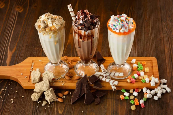 Three sweet milkshakes with chocolate, marshmallow, halva and whipped cream at a wooden board on table background.