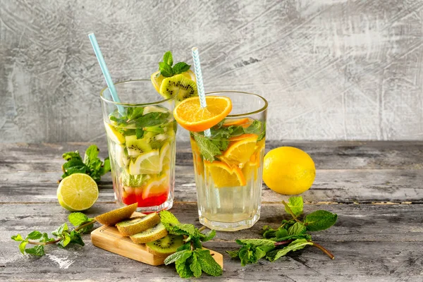 Two glasses of detox water with lemon, kiwi, orange and mint at concrete decorated table background. Concept of healthy lifestyle.