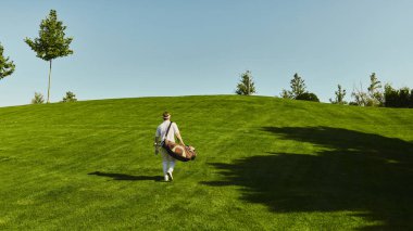 Golf oyuncusu güneşli sahada golf çantasıyla yokuş yukarı yürüyor. Aktif yaşam tarzı, seyahat eğlencesi, spor tutkusu, zihinsel denge ve açık hava sağlığı ilhamı kavramı.