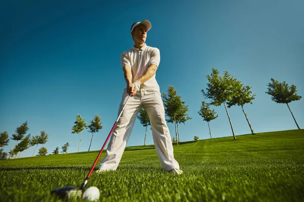 Golfçü mavi gökyüzünün altında çimlerin üzerinde topla atış yapmaya hazırlanıyor. Odaklanma kavramı, açık hava eğitimi, doğruluk, aktif yaşam tarzı ve spor adanmışlığı..